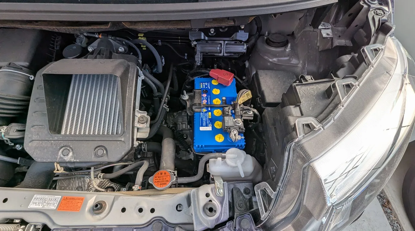 紫色のトヨタ ルーミーの開けられたエンジンルームのクローズアップ。中央には新しく換装されたパナソニック製の鮮やかな青いカオスバッテリー(N-80)が設置されている。